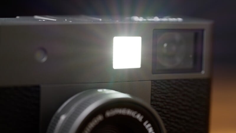 Close-up of a camera with its flash firing, emitting a bright burst of light. The lens and textured body are visible, with the flash creating a starburst effect in the image.