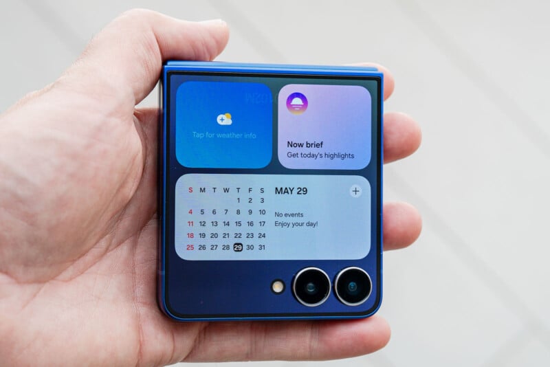 A hand holds a purple flip smartphone showing its external screen with widgets displaying weather info, calendar, and highlights for May 29. Two camera lenses are visible at the bottom right of the phone.