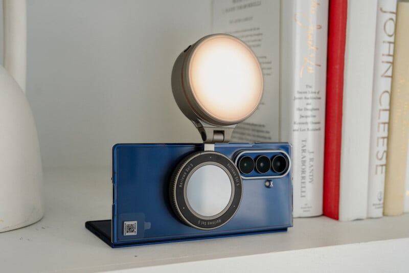 A blue smartphone with a circular camera lens attachment and a large round light mounted on top sits on a white shelf beside upright books.