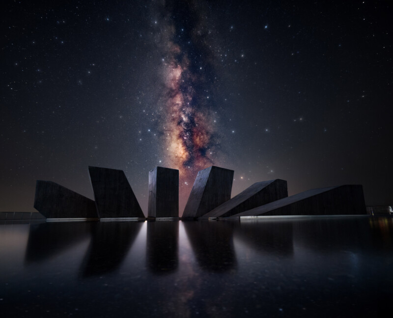 A dramatic night sky filled with stars and the glowing Milky Way above angular, dark stone structures reflected in calm water, creating a surreal and striking scene.