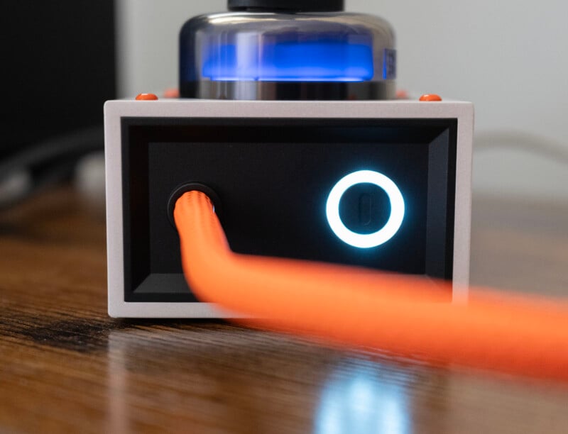 A close-up of an electronic device with a glowing blue circular power button, an orange braided cable plugged in, and a glass vacuum tube on top, all placed on a wooden surface.