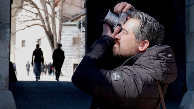 Man in a black jacket takes a photo in a shadowed area with a camera. Sunlit street and blurred figures are in the background.