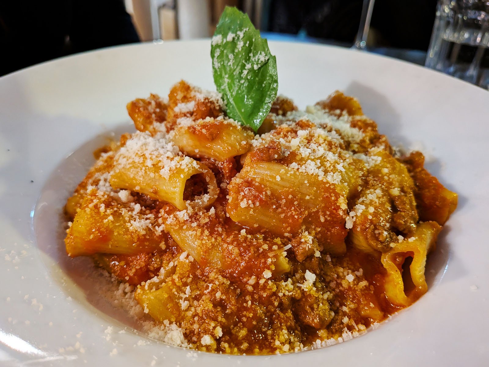 A white plate of rigatoni pasta with meat sauce, topped with grated cheese and garnished with a fresh basil leaf.