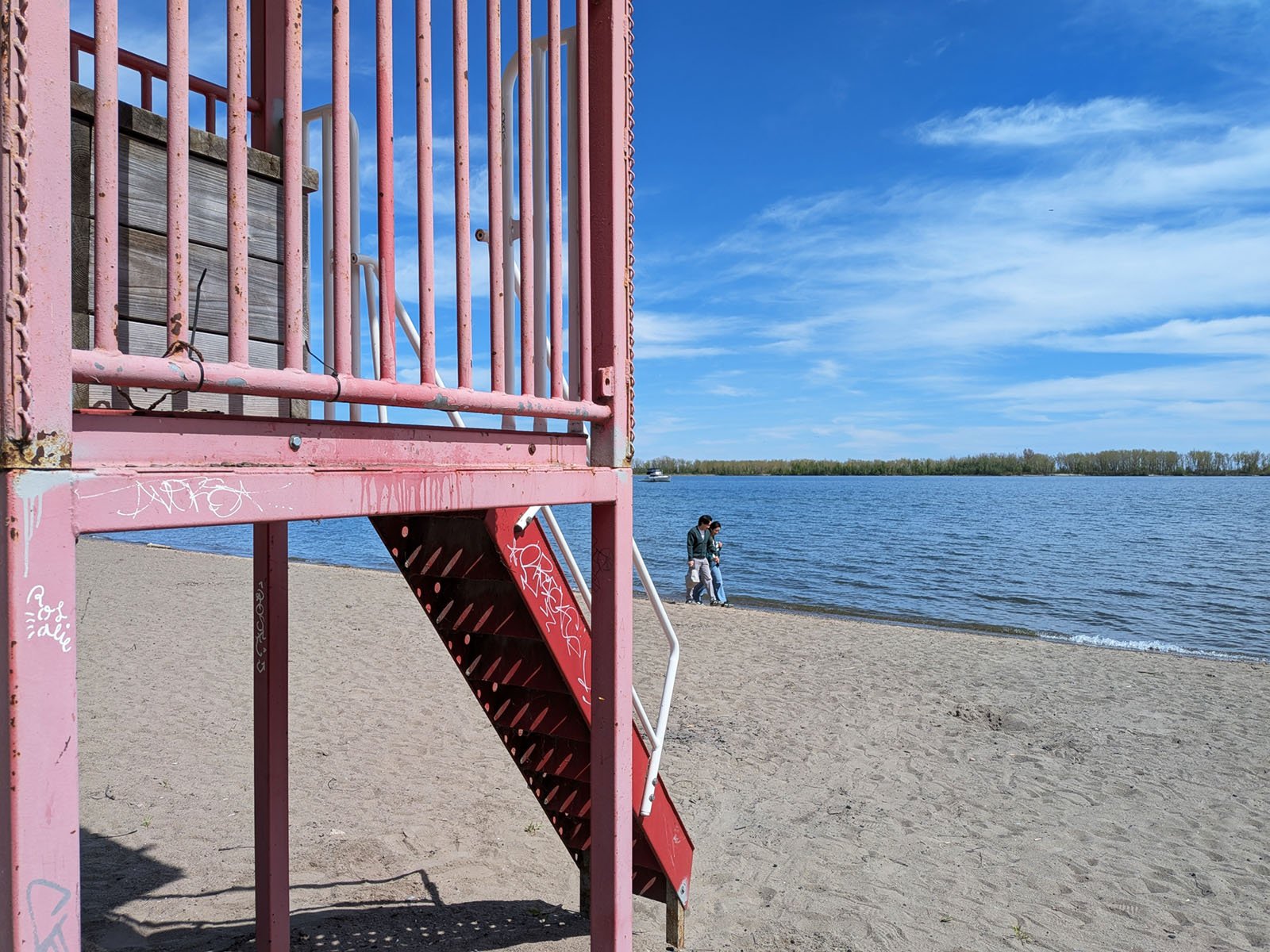 A pink lifeguard tower stands on a sandy beach beside a calm lake under a blue sky. Two people walk by the water's edge in the distance. Some graffiti is visible on the tower.