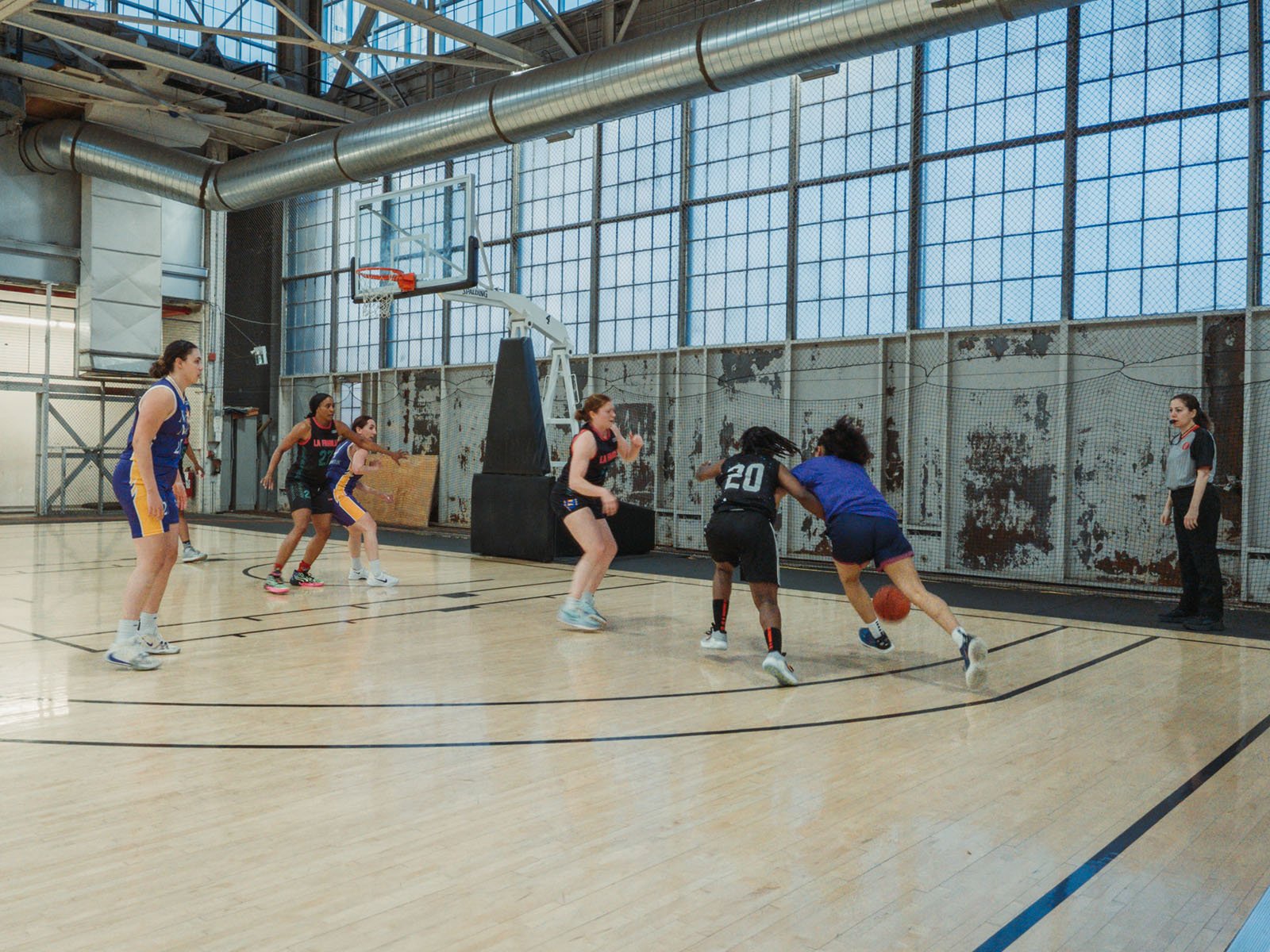 Five women play basketball indoors on a court; three wear purple and yellow uniforms while two wear black. A referee stands at the sideline, observing the game. The scene takes place in a gym with large windows.