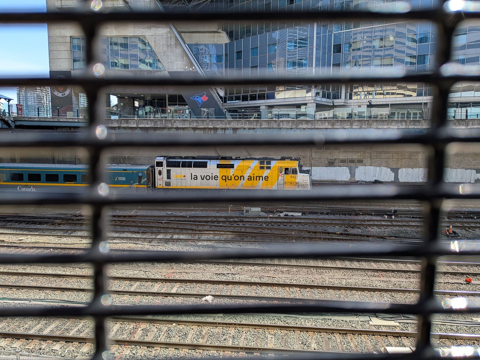 A yellow and white VIA Rail train with "la voie qu'on aime" on its side is seen through a metal fence, with railway tracks and a modern glass building in the background.