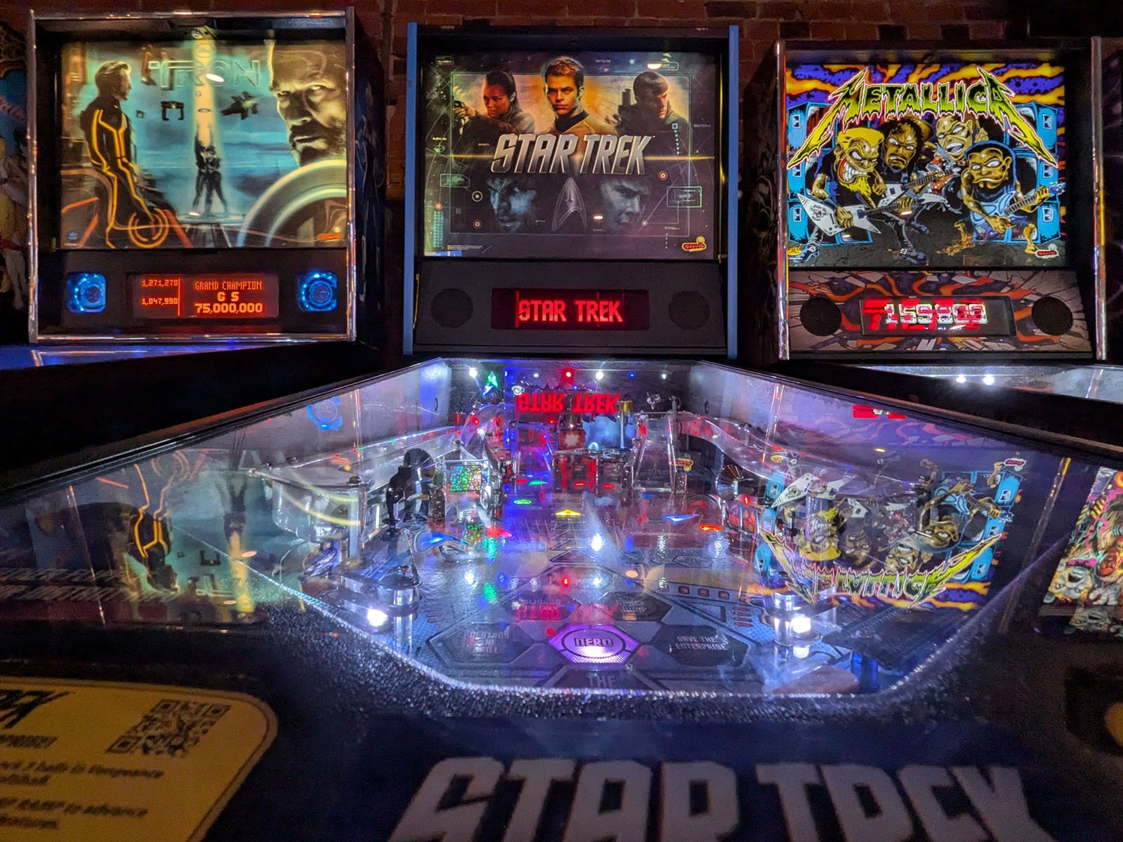 A brightly lit Star Trek pinball machine stands between Tron and Metallica pinball machines, with colorful lights reflecting off the glass and vibrant sci-fi themed artwork on each backboard.
