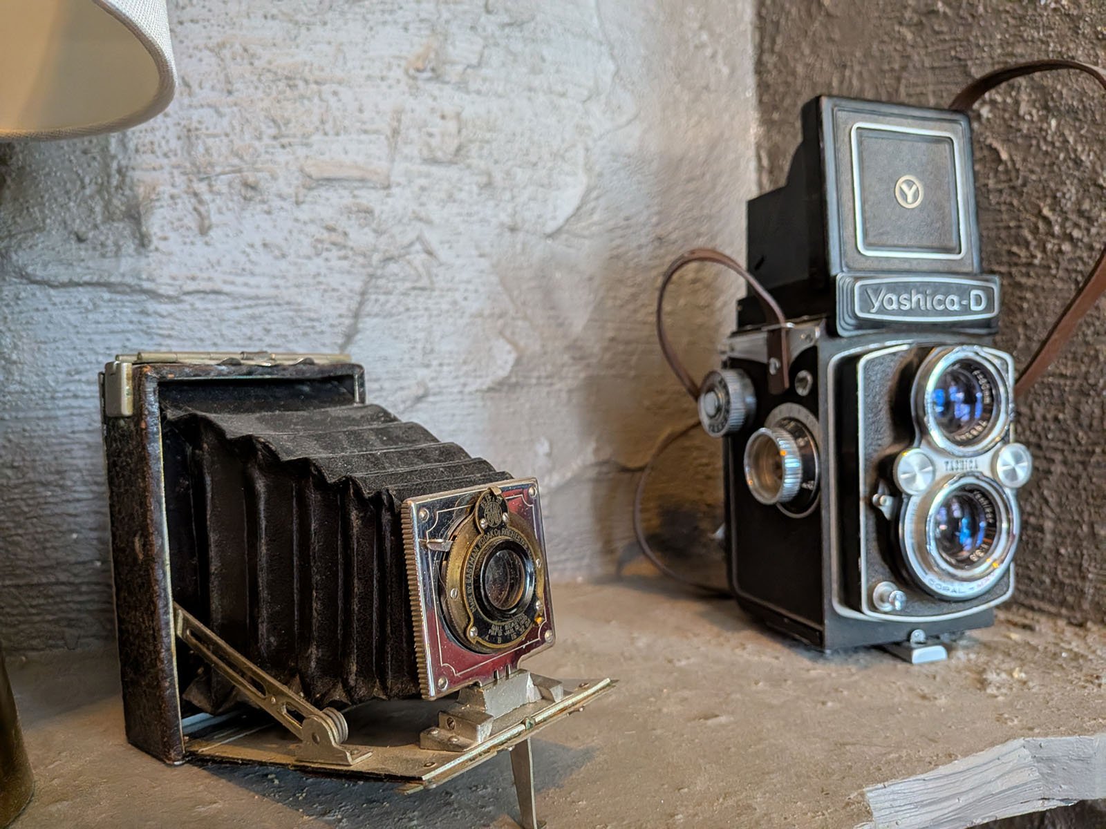 Two vintage cameras on a textured surface: a black bellows camera with brass details on the left, and a black Yashica-D twin-lens reflex camera with blue-tinted lenses on the right.