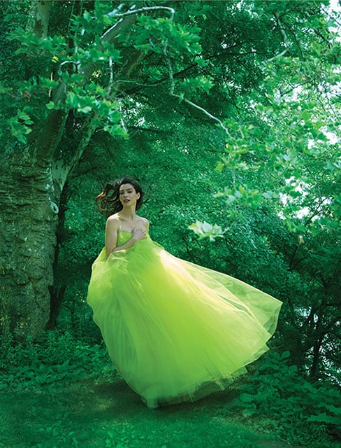 A woman in a flowing yellow-green gown stands gracefully in a lush, green forest, surrounded by trees and foliage. Sunlight filters through the leaves, creating a dreamy, ethereal atmosphere.