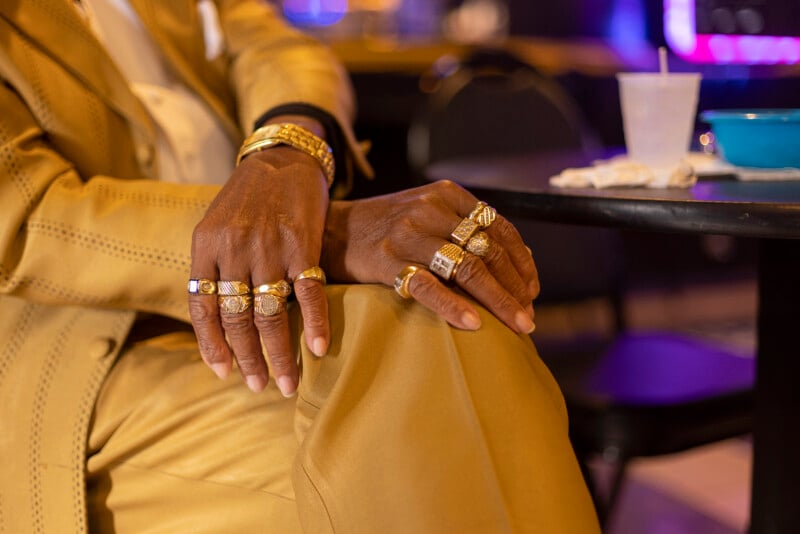 A person in a gold suit sits with legs crossed, displaying multiple gold rings and a gold watch on their hands. A table with a drink and napkins is visible in the background.