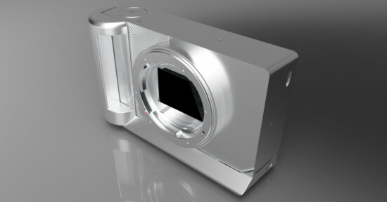 A silver, rectangular digital camera body without a lens is shown on a grey surface, featuring a prominent grip and a large circular lens mount in the center.