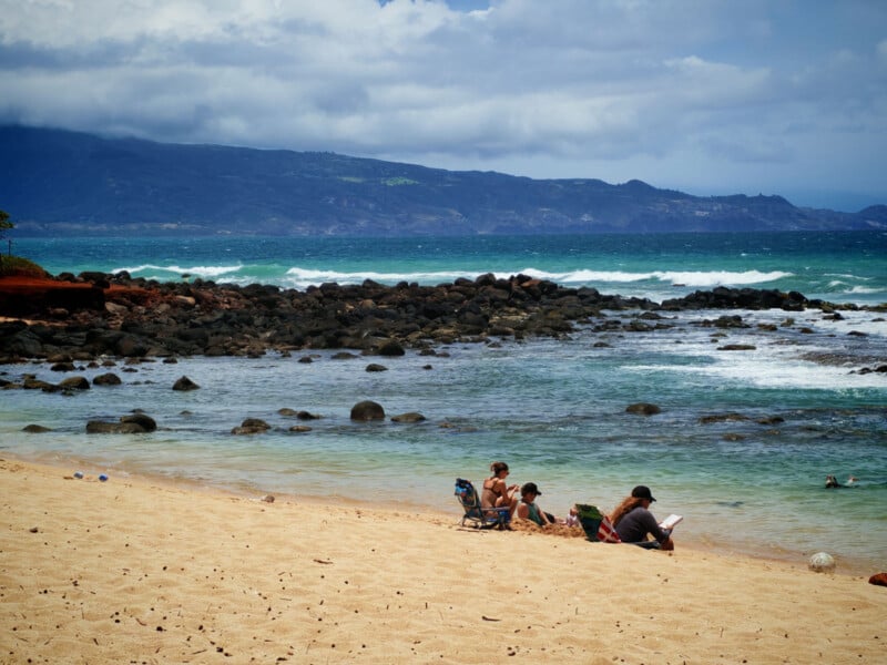 People relax on sandy beach near rocky shoreline with turquoise waves. Mountains and cloudy sky are visible in the background. Some people sit on chairs, while others play near the water.