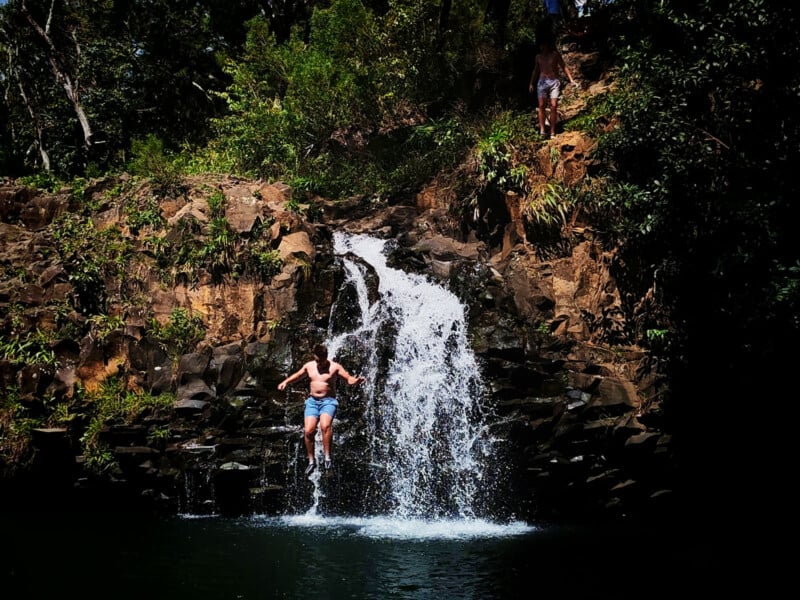 A person in swimwear leaps from a rocky ledge into a waterfall pool, surrounded by lush green foliage and trees, with another person standing above the waterfall.
