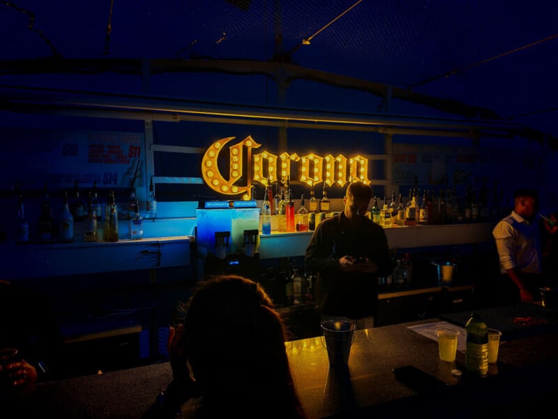 A dimly lit bar with a glowing "Corona" sign behind the counter. Bottles and glasses line the shelves. Two bartenders stand behind the bar, while a few customers face them in the foreground.