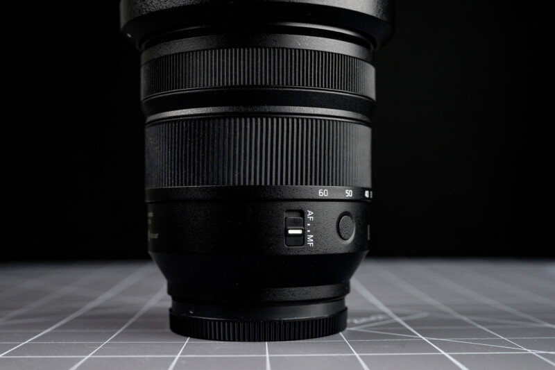 A close-up of a black camera lens with a textured focus ring, an AF/MF switch, and focal length markings, placed on a gray grid-patterned surface against a dark background.