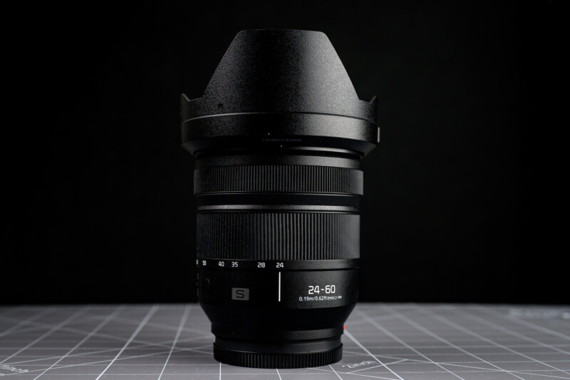 A black camera lens with a lens hood, labeled "24-60mm," standing upright on a grid-patterned gray surface against a dark background.