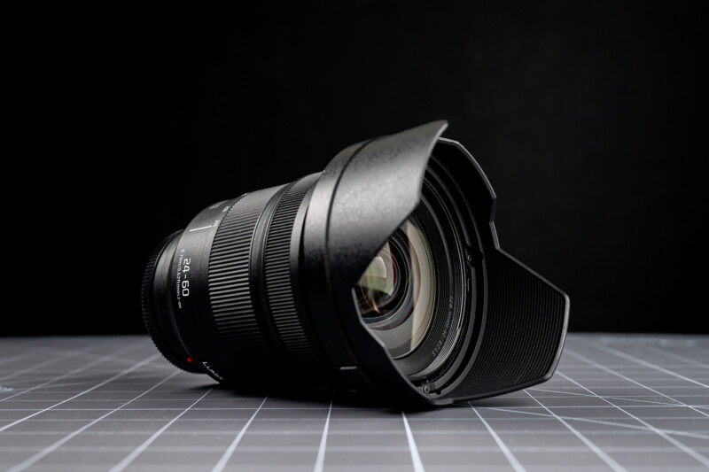 A black camera lens with a lens hood rests on a gray grid-patterned surface, set against a dark background.