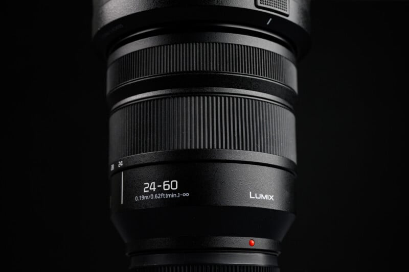 Close-up photo of a black Lumix camera lens with a 24-60mm zoom range, showing detailed texture and markings, set against a dark background.