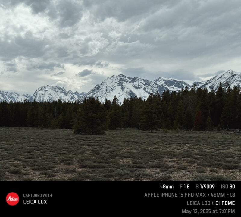 A cloudy sky hangs over snow-capped mountains and a dense forest, with a grassy field in the foreground. Photo details and Leica branding are overlaid at the bottom.