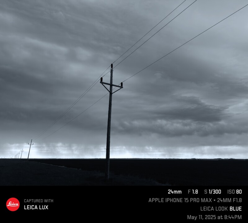 A moody, overcast sky stretches above a flat landscape with power lines receding into the distance. The scene is tinted blue, creating a dramatic and somber atmosphere.