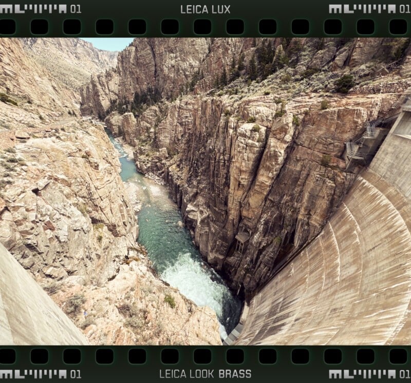 A dramatic view from the top of a large concrete dam, looking down at a narrow river winding through steep rocky cliffs, with sparse green vegetation scattered on the rocks.