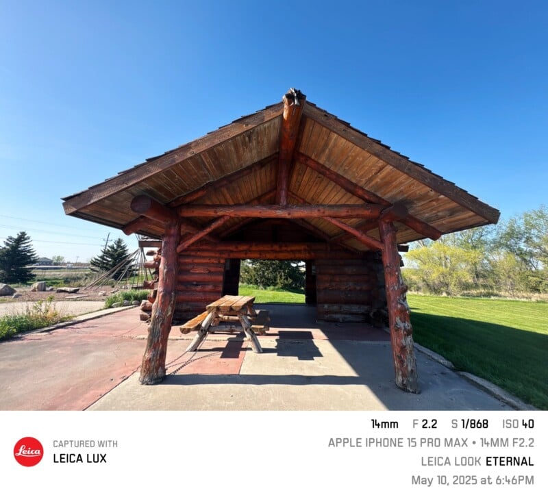 A rustic wooden pavilion with picnic tables is set in a park under a clear blue sky, surrounded by green trees and grass. Photo taken with an iPhone 15 Pro Max on May 10, 2025.