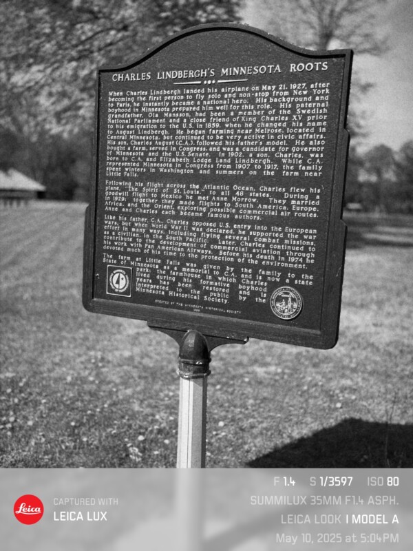A black-and-white photo of a historical marker titled "Charles Lindbergh's Minnesota Roots," with text about Lindbergh. The marker stands on a grassy lawn. Camera details are displayed at the bottom.