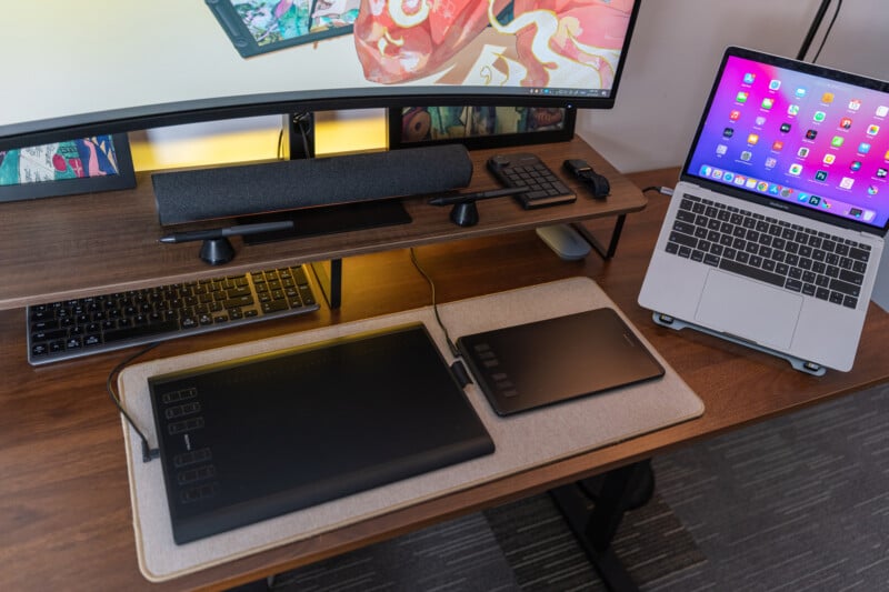 A modern desk setup with a large curved monitor, a laptop on a stand, wireless keyboard and mouse, drawing tablets, and a soundbar, all arranged neatly on a wooden desk with an organized workspace.