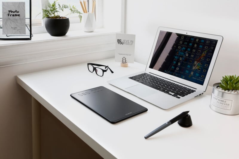 A tidy white desk with a laptop, drawing tablet, stylus, glasses, potted plants, a “Believe in Yourself” card, and a photo frame near a bright window.