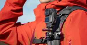 A person wearing a red jacket has a black action camera mounted on the strap of their black backpack, ready for hands-free recording outdoors.