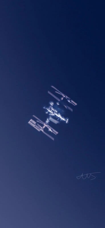 The International Space Station orbits against a deep blue sky, its solar panels and modules clearly visible as sunlight reflects off its structure.