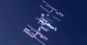 A blurred, close-up image of the International Space Station (ISS) orbiting against a dark blue sky, with its solar panels and structural modules visible.