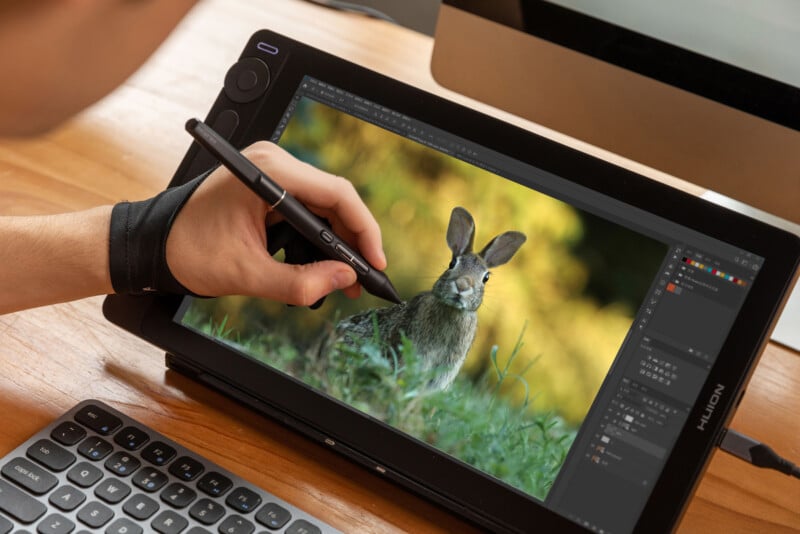 A person uses a stylus on a Huion drawing tablet to edit a photo of a rabbit in Photoshop, with a keyboard and monitor visible on a wooden desk.