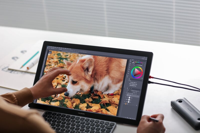 A person edits a photo of a corgi lying on autumn leaves using a digital drawing tablet and stylus at a desk, with art supplies nearby.