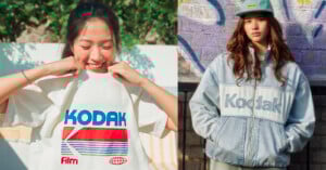 Two young women wear Kodak-branded clothing; one smiles while pulling up a white Kodak T-shirt, and the other stands against a wall in a light denim Kodak jacket and cap.