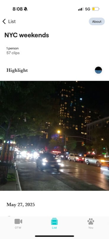Nighttime city street in NYC filled with cars and bright headlights, with tall buildings illuminated in the background. The sky is dark, and trees line the sidewalk.