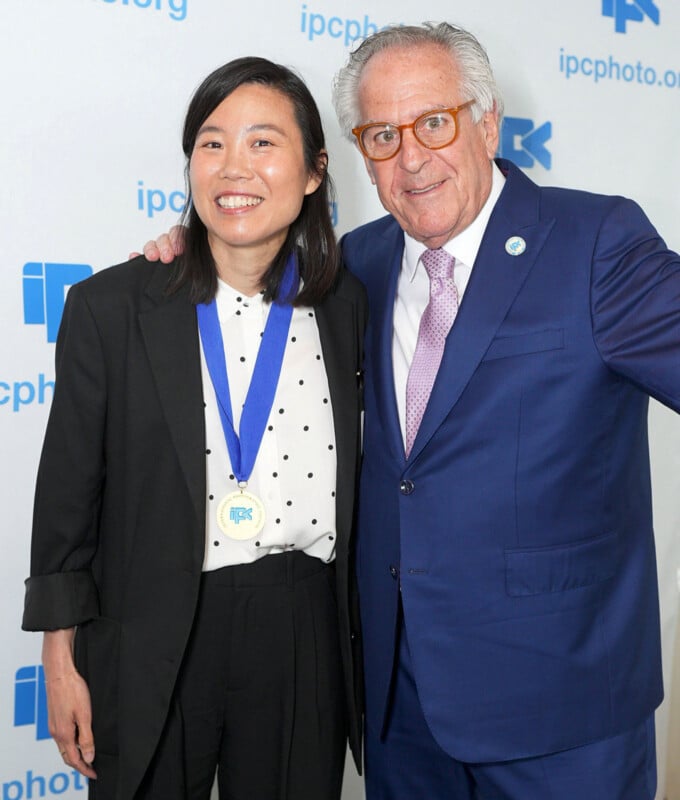 Two people stand together smiling at an event. The woman on the left wears a black suit, white shirt with black dots, and a medal. The man on the right wears a blue suit and glasses. The backdrop displays "ipcphoto.org" and blue logos.