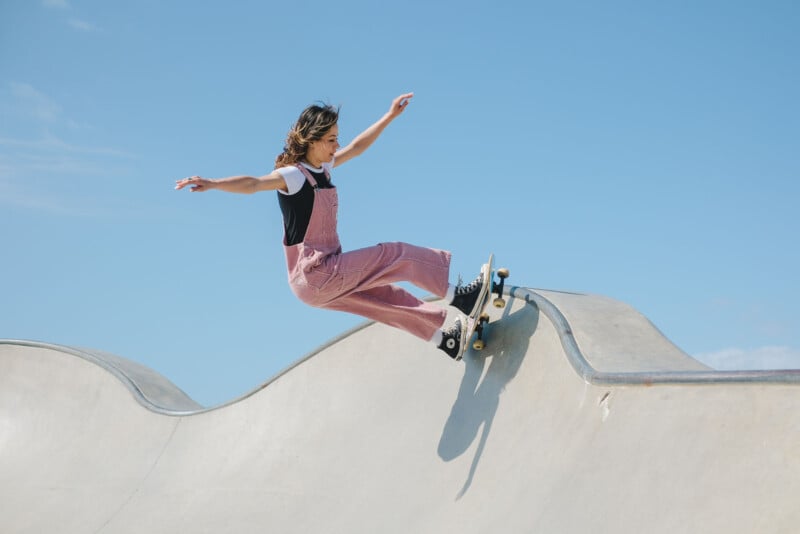 A person in pink overalls and a white t-shirt is skateboarding on the curved edge of a concrete skatepark under a clear blue sky. They have their arms outstretched for balance.