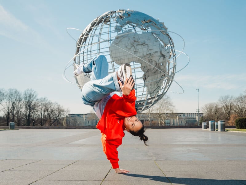 A person in a red jacket performs a one-handed breakdance move on a plaza, with a large metal globe sculpture in the background under a clear blue sky.