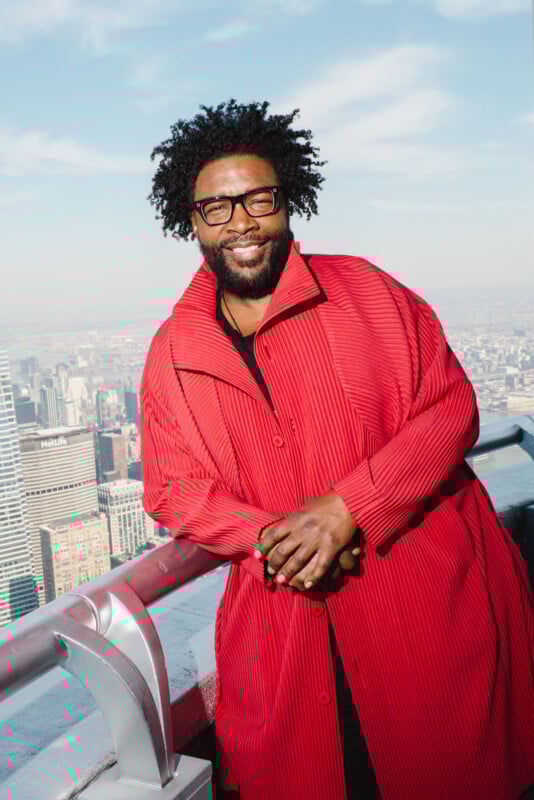 A man with curly hair and glasses smiles while leaning on a railing atop a city skyscraper, wearing a bright red textured coat, with the cityscape visible in the background under a blue sky.