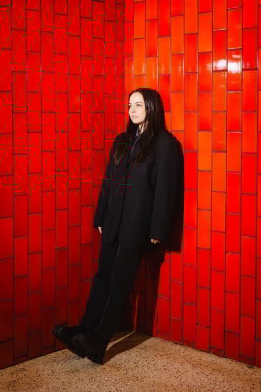 A person with long dark hair, wearing a black suit, stands against a corner of glossy red tiled walls on a speckled beige floor, looking forward with a neutral expression.