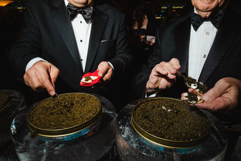 Two people in tuxedos serve black caviar onto small food items from large tins at a formal event. Only their hands and torsos are visible. The background is dark and out of focus.