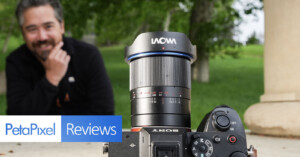 A camera with a Laowa lens is in focus in the foreground, with a smiling man sitting in the blurred background. The PetaPixel Reviews logo appears at the bottom left. The scene is outdoors with green trees and grass.