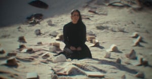 A woman in black sits cross-legged on a blanket in the sand, surrounded by open books and large stones scattered around her under bright daylight.