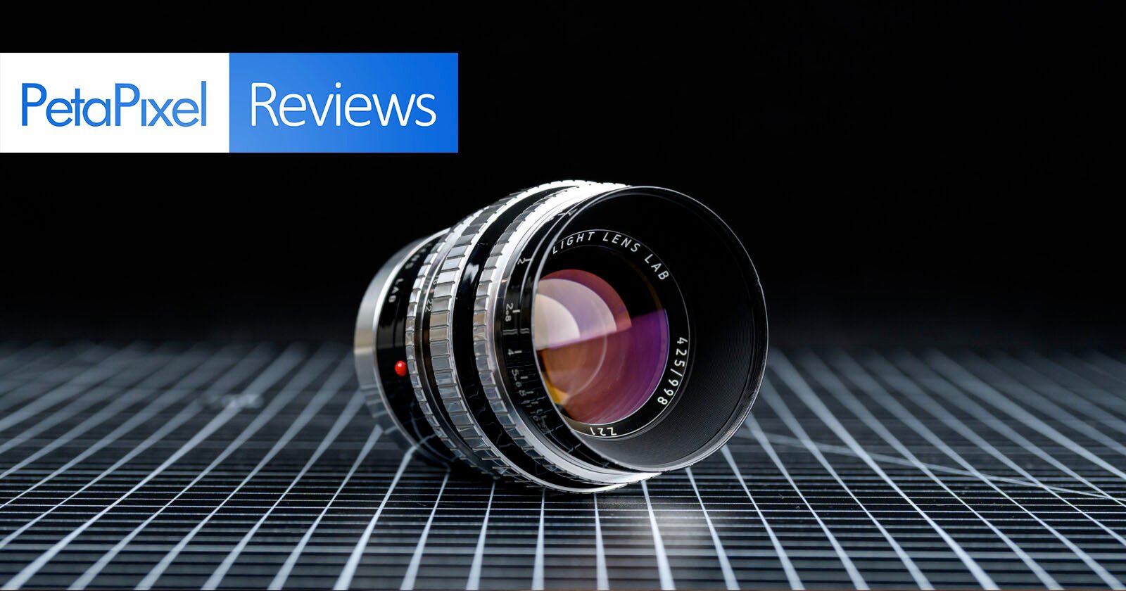 A camera lens rests on a grid-patterned surface against a black background. In the top left corner, the PetaPixel Reviews logo is displayed with blue and white text.