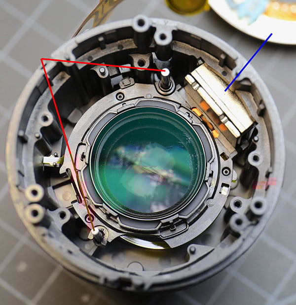 Close-up of a disassembled camera lens with visible internal components, glass elements, and electronic parts. Red and blue lines are drawn to highlight specific areas inside the lens housing.