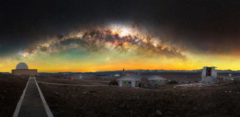 Panoramic view of an observatory under a vivid, star-filled night sky with the Milky Way arched overhead, glowing above buildings and a rocky, open landscape.