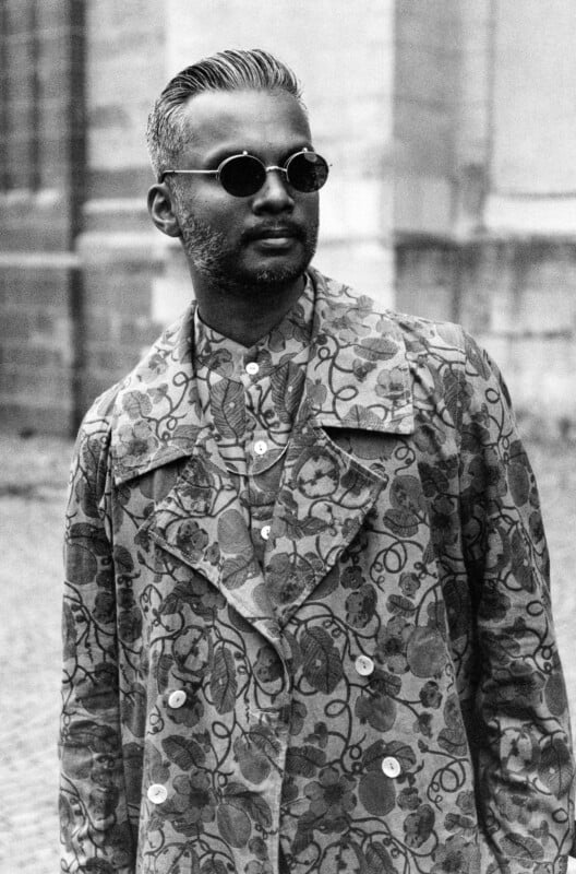 A man with short, gray hair and a beard wears round sunglasses and a patterned floral coat over a patterned shirt, standing outdoors in front of a stone building. The image is black and white.