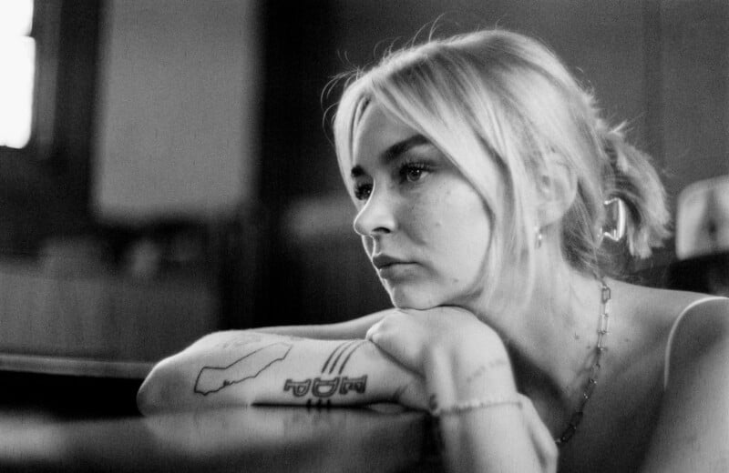 A black and white photo of a young woman with light hair resting her chin on her crossed arms, looking thoughtfully into the distance. She has tattoos on her forearm and wears a chain necklace.