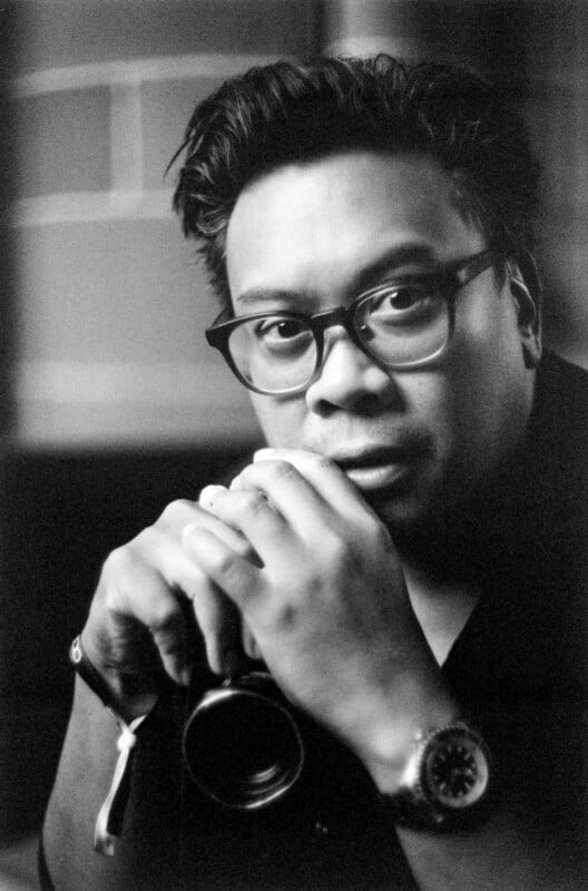 Black and white portrait of a person with short, dark hair and glasses, holding a camera close to their face with both hands, looking directly at the camera. The background is softly blurred.